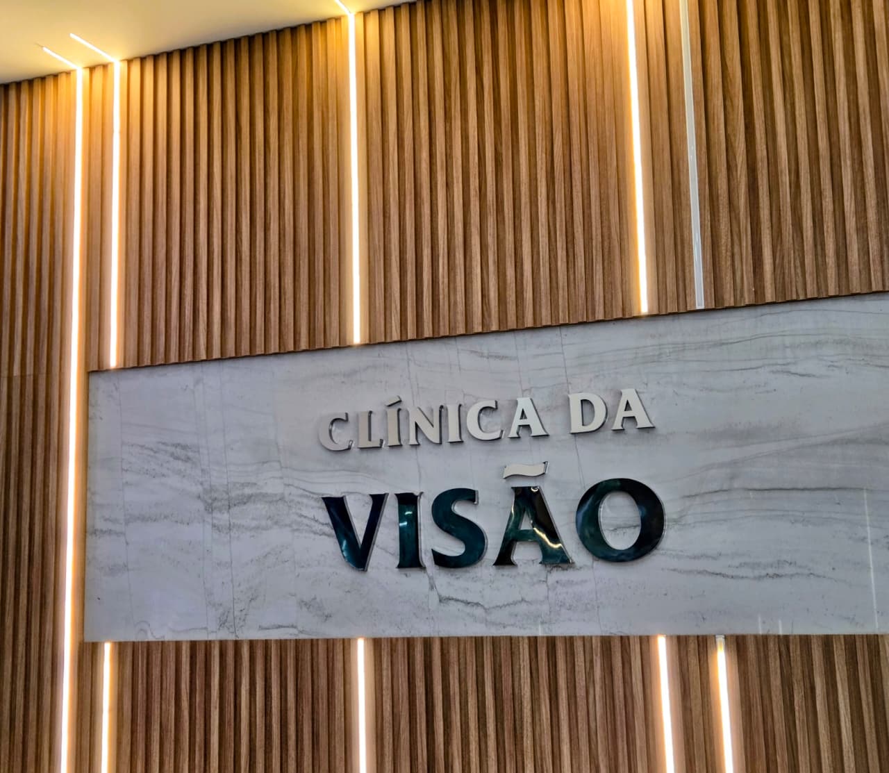 Letreiro da Clínica da Visão em mármore com iluminação âmbar sobre painel de madeira ripada
